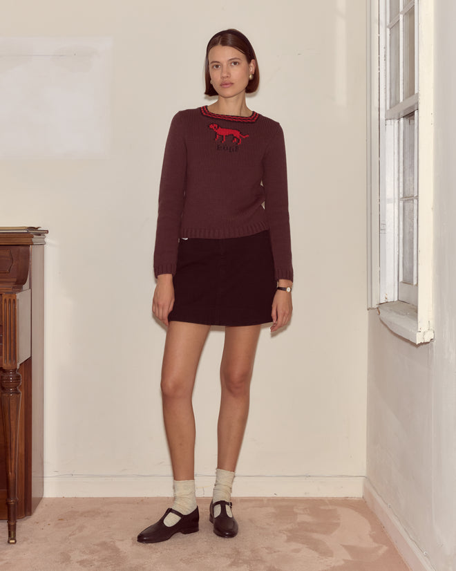 A woman stands indoors by a window, wearing the Lapdog Sweater in brown, paired with a black skirt, socks, and black shoes.