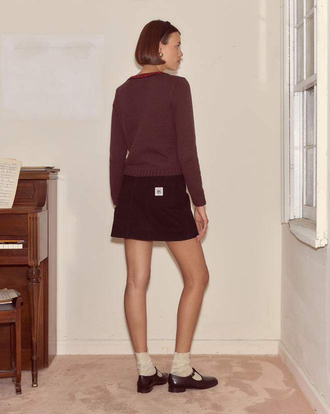 A woman wearing the Lapdog Sweater stands facing away in a room with knitwear accents, next to a piano and window, paired with a black skirt.