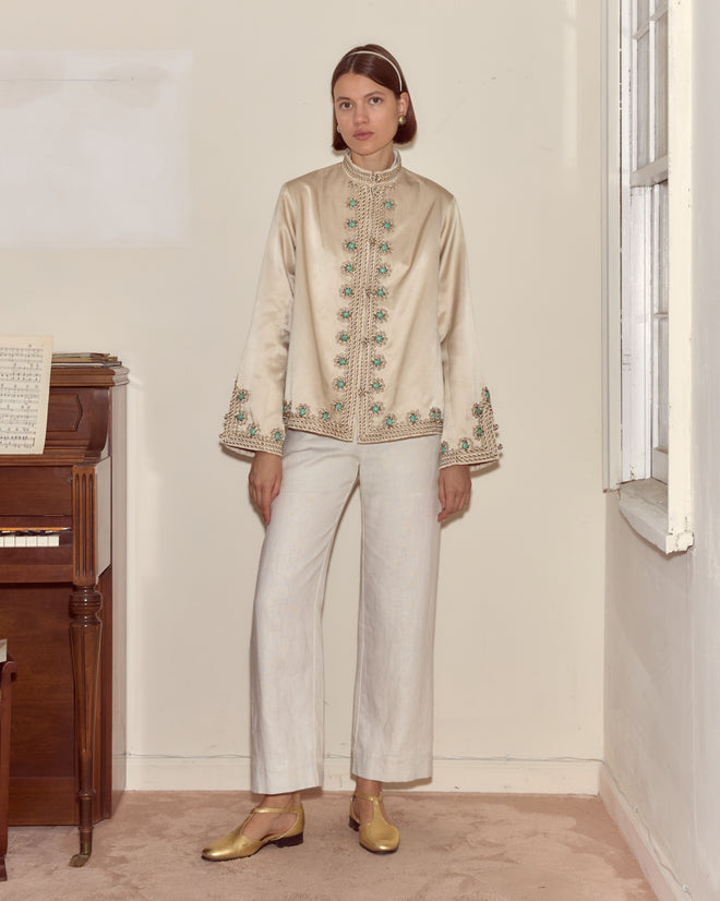 A woman wearing the Lapis Braid Jacket stands by a piano in a softly lit room with a window.