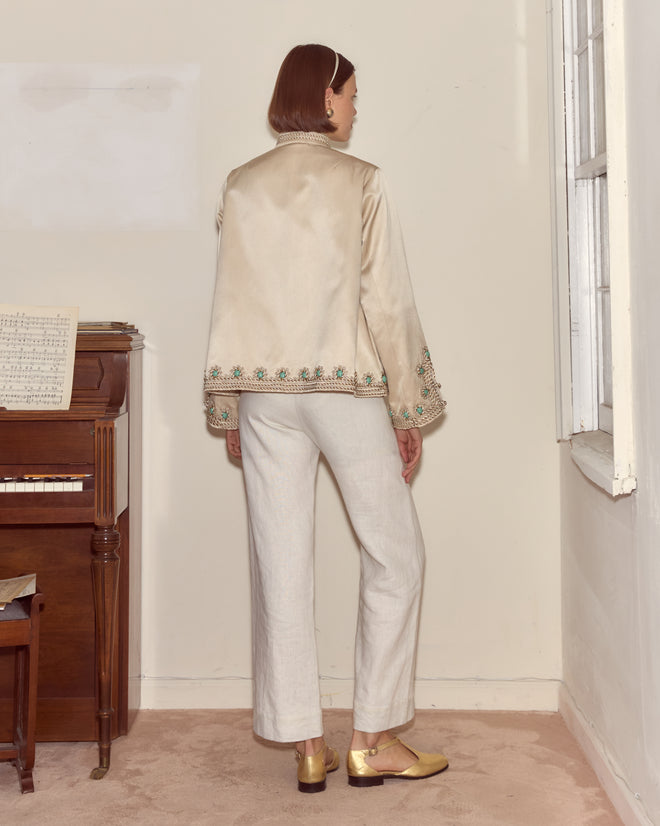 A person in the Lapis Braid Jacket and white pants stands with their back to the camera near a piano and window in a beige room.