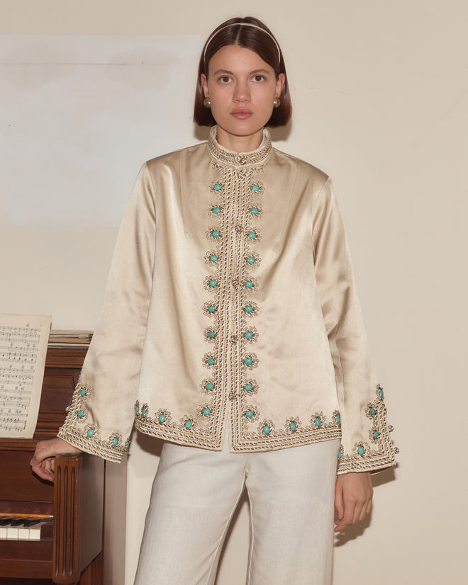 A woman stands beside a piano adorned with sheet music, wearing the Lapis Braid Jacket—a beige 1960s-inspired piece featuring ornate braided trim and turquoise accents.