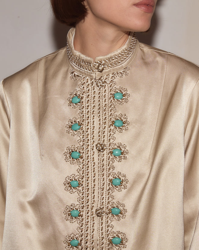 Person wearing the Lapis Braid Jacket, a cream satin 1960s-style coat featuring ornate gold braided trim and turquoise decorative buttons.