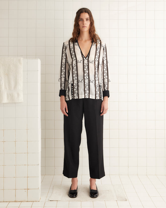 A person in the Lattice Sprig Top, black pants, and black shoes stands in a tiled room.