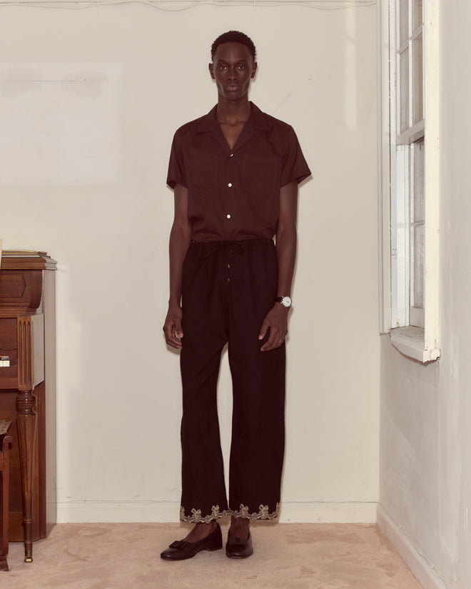 A person wearing Laurelist Pajama Pants - Black stands indoors near a piano and a window, facing the camera.