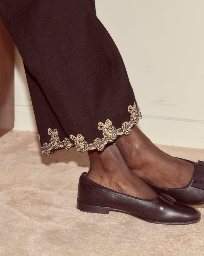 Close-up of a person's legs wearing Laurelist Pajama Pants - Black with vintage passementerie gold trim and black loafers on a beige carpet.