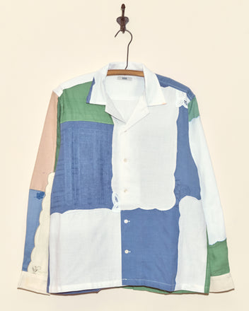 The Lavender Fields Napkin Shirt - M/L, a unique long-sleeve top with multicolored patchwork, hangs on a hook against a plain beige wall.