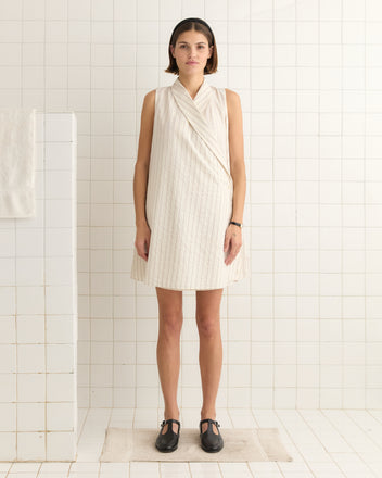 The woman in a white dress with black shoes stands in a tiled room, facing forward. Her Lavender Sail Dress, made from 100% cotton, includes antique pearl buttons that evoke the elegance of 1930s shirting.