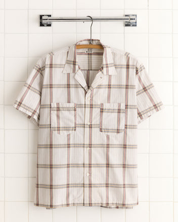 The Lawn Fence Shirt - XXL is a short-sleeve, button-up shirt crafted from unique deadstock plaid fabric, featuring two front pockets. Displayed on a metal hanger against white tiles.