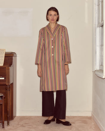 A woman in the Lawrence Coat with antique pearl buttons and black pants stands indoors beside a piano and a window.