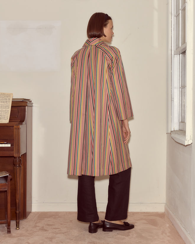 A woman in a formal Lawrence Coat of heavyweight fabric stands indoors near a window and piano, facing away from the camera.