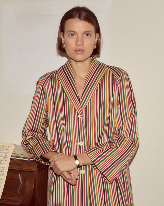 A woman wearing the Lawrence Coat—a formal 1950s-inspired design in heavyweight fabric with antique pearl buttons—stands by a piano with sheet music, looking at the camera.