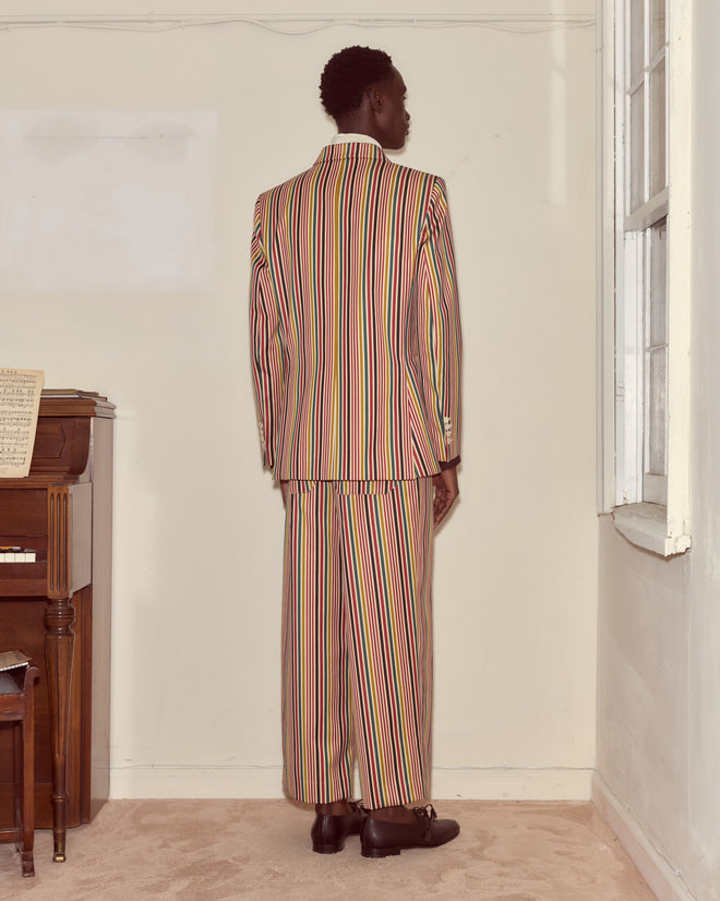 Wearing the Lawrence Suit Jacket—a colorful, single-breasted wool blazer with stripe details and antique pearl buttons—a person stands facing a wall near a piano and window in a carpeted room.
