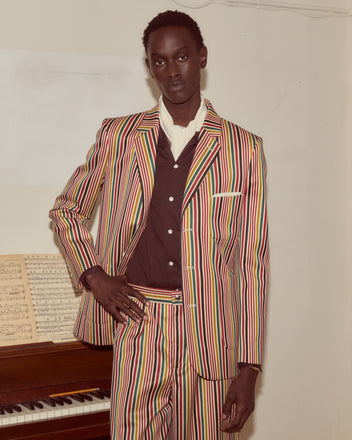 Wearing the Lawrence Suit Jacket, a person stands confidently by a piano with sheet music, showcasing the colorful single-breasted striped wool design and antique pearl buttons.