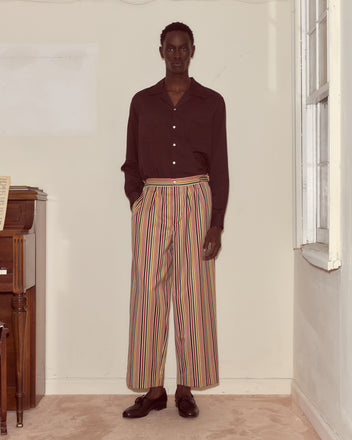 Indoors, a person stands next to a piano wearing a dark shirt and the Lawrence Trousers—wide-leg wool trousers featuring colorful stripes and side-buckle waist adjusters.