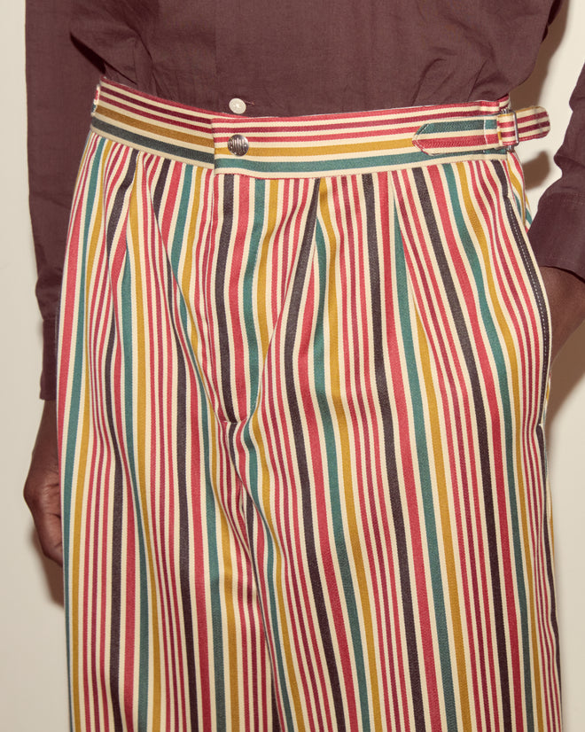 Person wearing the Lawrence Trousers—colorful, vertically striped wool pants with a wide-leg silhouette—and a brown shirt, hand by their side.