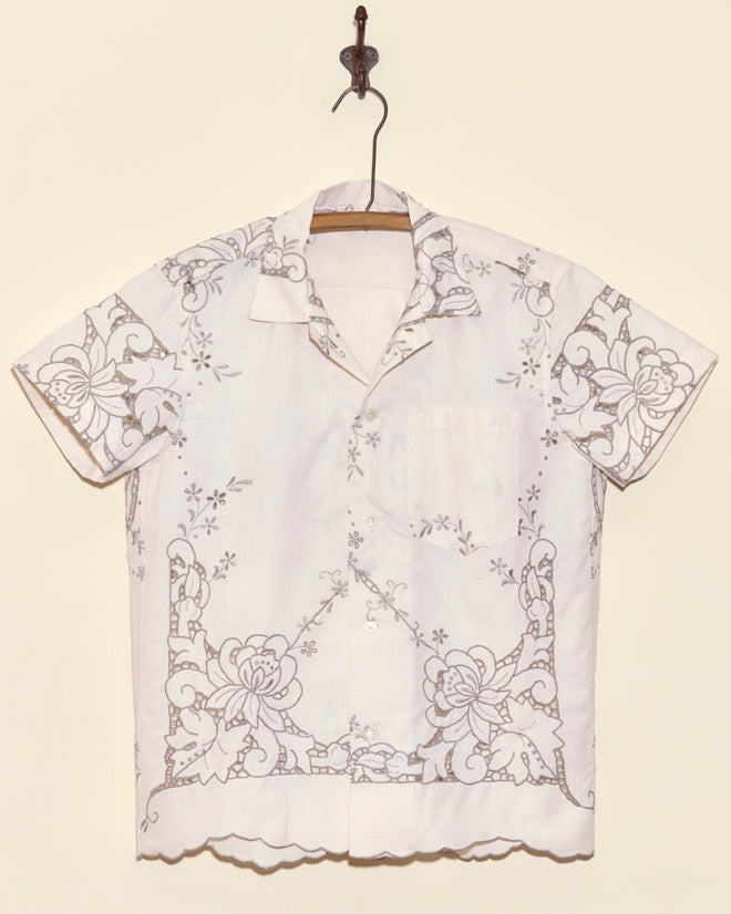 Layered Iris Short Sleeve Shirt in XS, featuring white fabric with intricate floral cutwork embroidery, displayed on a hanger against a beige wall.