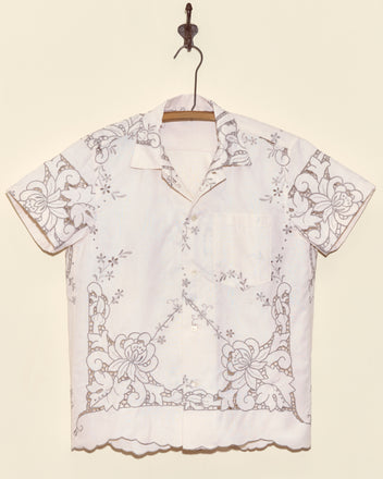 Layered Iris Short Sleeve Shirt in XS, featuring white fabric with intricate floral cutwork embroidery, displayed on a hanger against a beige wall.