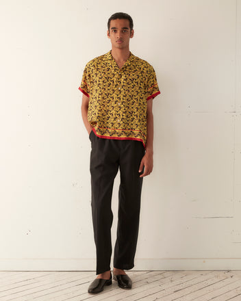 A person stands against a white wall wearing a Lemon Paisley Short Sleeve Shirt, black pants, and black shoes. They have their hands in their pockets and look directly at the camera.