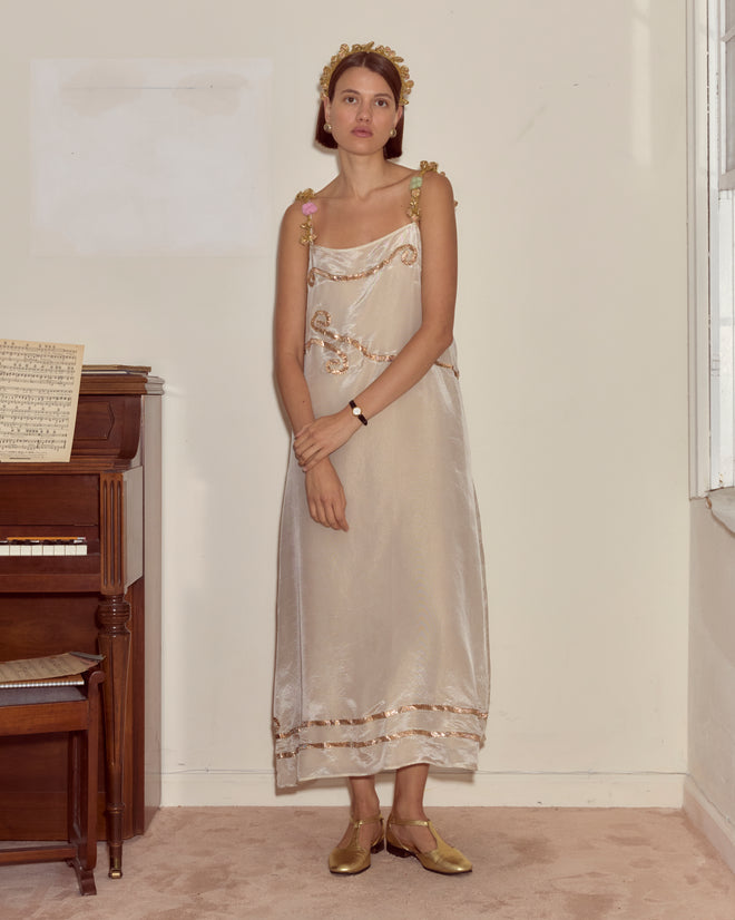 Woman in the Lido Dress, featuring gold trim and cream appliqué, stands by a piano, accessorized with a gold headband and gold shoes.