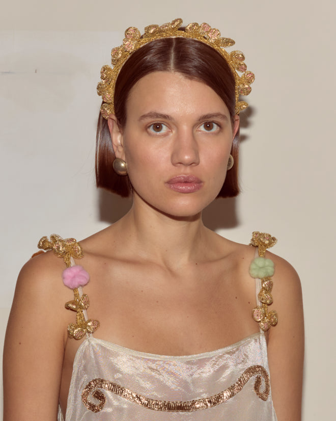 Woman with straight brown hair wears the Lido Headband, a gold Peter Pan style with lamé leaves, paired with pearl earrings and a decorated white dress.