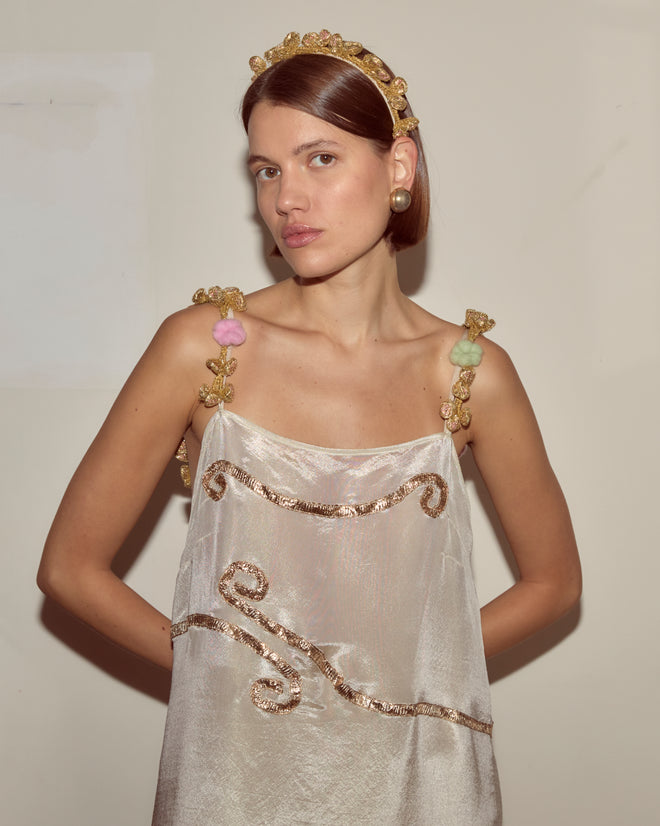 A woman in the Lido Dress, a shimmering white appliqué gown with gold accents, paired with a gold headband, stands against a plain background.