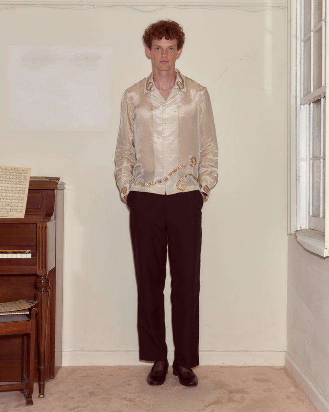 Wearing the Lido Long Sleeve and black pants, a person stands indoors by a piano and window, radiating refined elegance.