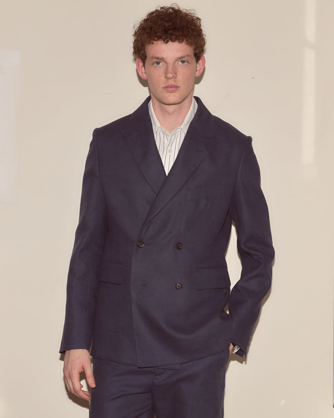 A young man with curly hair wears the Lightweight Linen Double-Breasted Suit Jacket in Midnight over a striped shirt, standing against a plain light-colored wall.