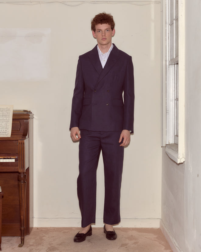 A man wearing the Lightweight Linen Double-Breasted Suit Jacket in Midnight stands indoors by a piano and a window, paired with black loafers.