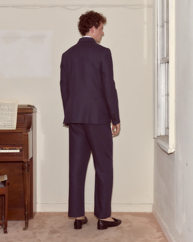 A man in the Lightweight Linen Double-Breasted Suit Jacket - Midnight stands near a piano and window, facing the corner of a room.