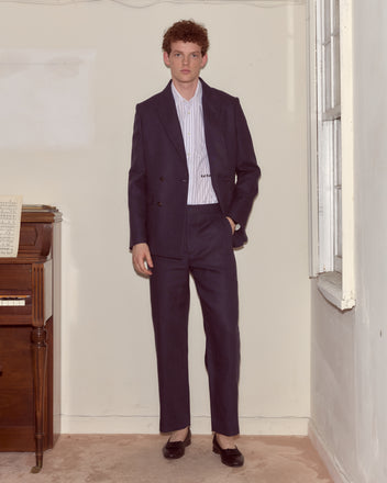 A man in the Lightweight Linen Double-Breasted Suit Jacket - Midnight stands indoors by a piano and window, facing the camera.
