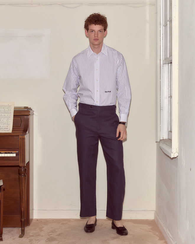 A man in a striped shirt and Lightweight Linen Side-Buckle Trousers - Midnight stands indoors beside a piano with sheet music.