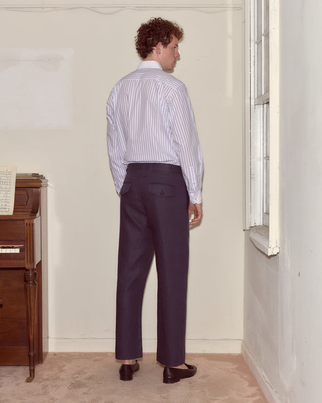 A man in a striped shirt and Lightweight Linen Side-Buckle Trousers - Midnight stands facing a wall near a piano and window.