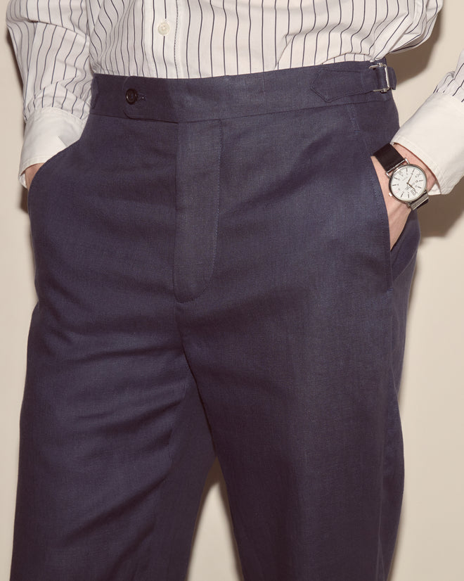 Man wearing Lightweight Linen Side-Buckle Trousers in Midnight, a striped white shirt, and a black wristwatch with hands in his pockets.