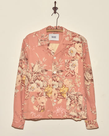 The Lily Rose Long Sleeve Shirt (XS/S), a light pink floral button-up with a collar, hangs on a hook against a plain wall, styled as a unique piece.