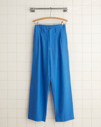 Linen Murray Trousers in Indigo, high-rise wide-leg style, hanging on a wooden hanger against a white tiled wall.