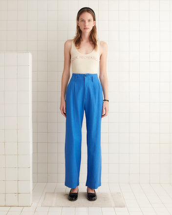 Linen Murray Trousers in Indigo, high-rise wide-leg style, hanging on a wooden hanger against a white tiled wall.