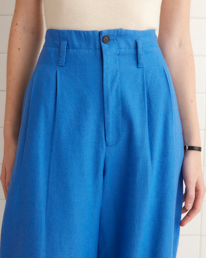 Linen Murray Trousers in Indigo, high-rise wide-leg style, hanging on a wooden hanger against a white tiled wall.