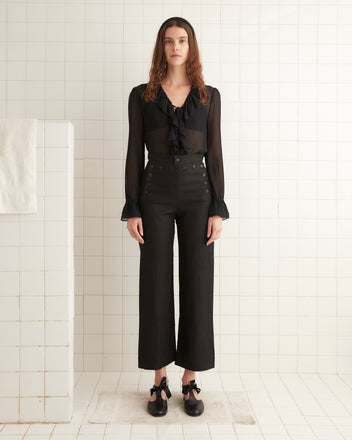 A woman stands in a tiled room wearing a black blouse and Linen Sailor Trousers - Black, which are high-waisted and 1940s-inspired with button details, complemented by black shoes with ankle ties.