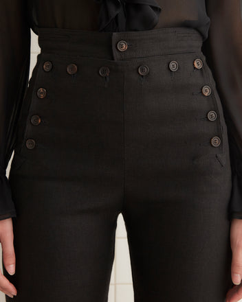 Someone is wearing Linen Sailor Trousers - Black with multiple buttons and a sheer black blouse, exuding style and confidence.