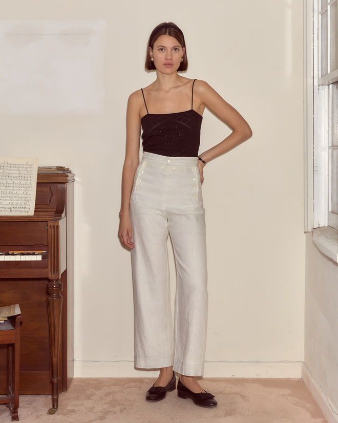 Woman in a black top and Linen Sailor Trousers - White stands beside a piano in a softly lit room.