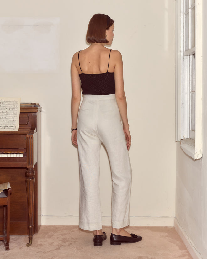 A woman stands indoors facing a wall, wearing a black top and Linen Sailor Trousers - White, next to a piano with sheet music.