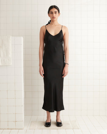 A person wearing the Lizzie Long Slip in black stands in a white tiled room, looking directly at the camera.