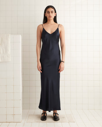 A woman wearing the Lizzie Long Slip in midnight satin stands gracefully in a white tiled room, exuding 1940s elegance.