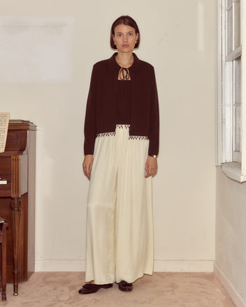 Woman standing indoors by a piano, wearing the Lodge Cardigan in Dark Brown layered under a black jacket with white wide-leg pants, illuminated by natural light.