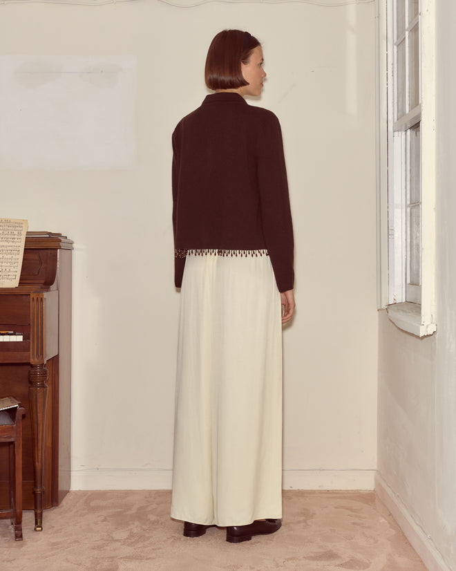 A woman stands indoors near a piano and window, facing a wall while wearing the Lodge Cardigan in Dark Brown and a long white skirt.