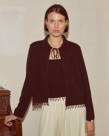 A woman wearing the Lodge Cardigan in dark brown and white pants stands indoors by a wooden piano, looking at the camera.