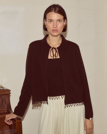 Woman with straight brown hair in a black top and white pants stands indoors by a wooden piano, layering her outfit with the Lodge Cardigan - Black.