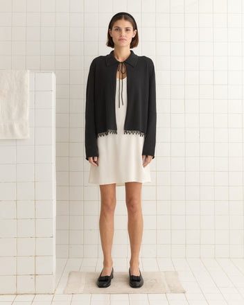 A woman stands in a tiled room wearing a Lodge Cardigan - Black over a white dress and black shoes on a beige mat.