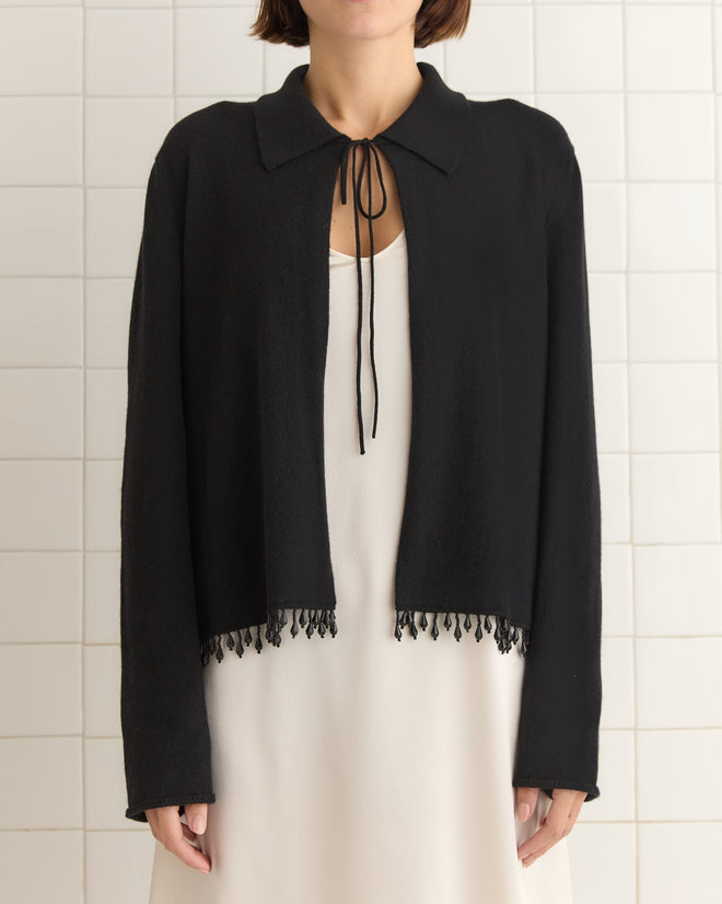 Elegantly dressed in a Lodge Cardigan in black over a white dress, the person stands against a tiled wall.