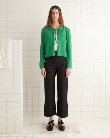 A person stands in a tiled room wearing a green Lodge Cardigan, white top, black pants, and black shoes.